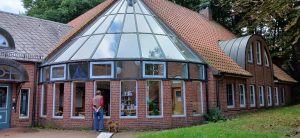 Ein Mann und ein Hund stehen vor einem Gewächshaus mit Glasdach und Backsteinwänden. Das Gebäude ist von Grünflächen umgeben.