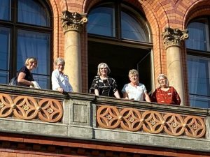 Fünf Frauen stehen auf einem Balkon mit ornamentalem Geländer vor einem Gebäude mit großen Fenstern und Säulen.