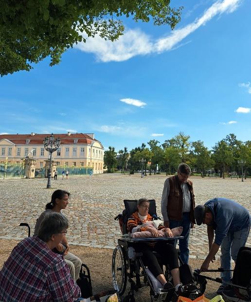 Eine Gruppe von Menschen, darunter einige im Rollstuhl, versammelt sich auf dem Kopfsteinpflaster vor dem Schloss Charlottenburg in Potsdam.
