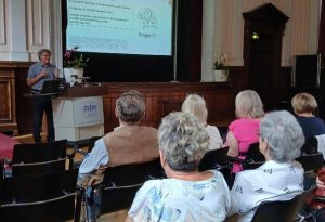 Eine Person hält einen Vortrag vor einem Publikum in einem Raum mit Holzvertäfelungen und einem Projektorbild im Hintergrund.
