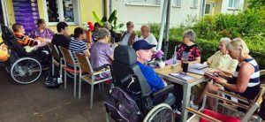 Eine Gruppe von Senioren, einige in Rollstühlen, trifft sich im Freien und genießt eine gesellige Runde mit Gesprächen und Getränken.