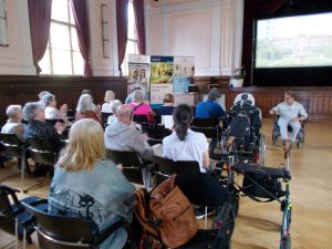 Eine Gruppe von Menschen, darunter einige im Rollstuhl, nimmt an einer Veranstaltung in einem Saal mit großen Fenstern und Vorhängen teil.