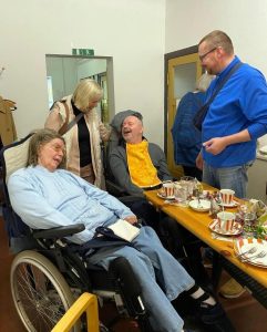 Eine Gruppe von Menschen teilt einen lustigen Momen. Eine Frau im Rollstuhl und ein Mann lachen, während andere ihnen zusehen.