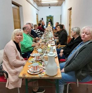 Eine Gruppe von Menschen sitzt an einem langen Tisch und genießt ein gemeinsames Essen. Der Tisch ist mit Tellern, Tassen und einer Milchkanne dekoriert.