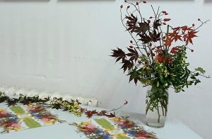 Ein Glas mit einem herbstlichen Blumenarrangement steht auf einem Tisch, daneben liegen verstreute Blätter und Blüten.