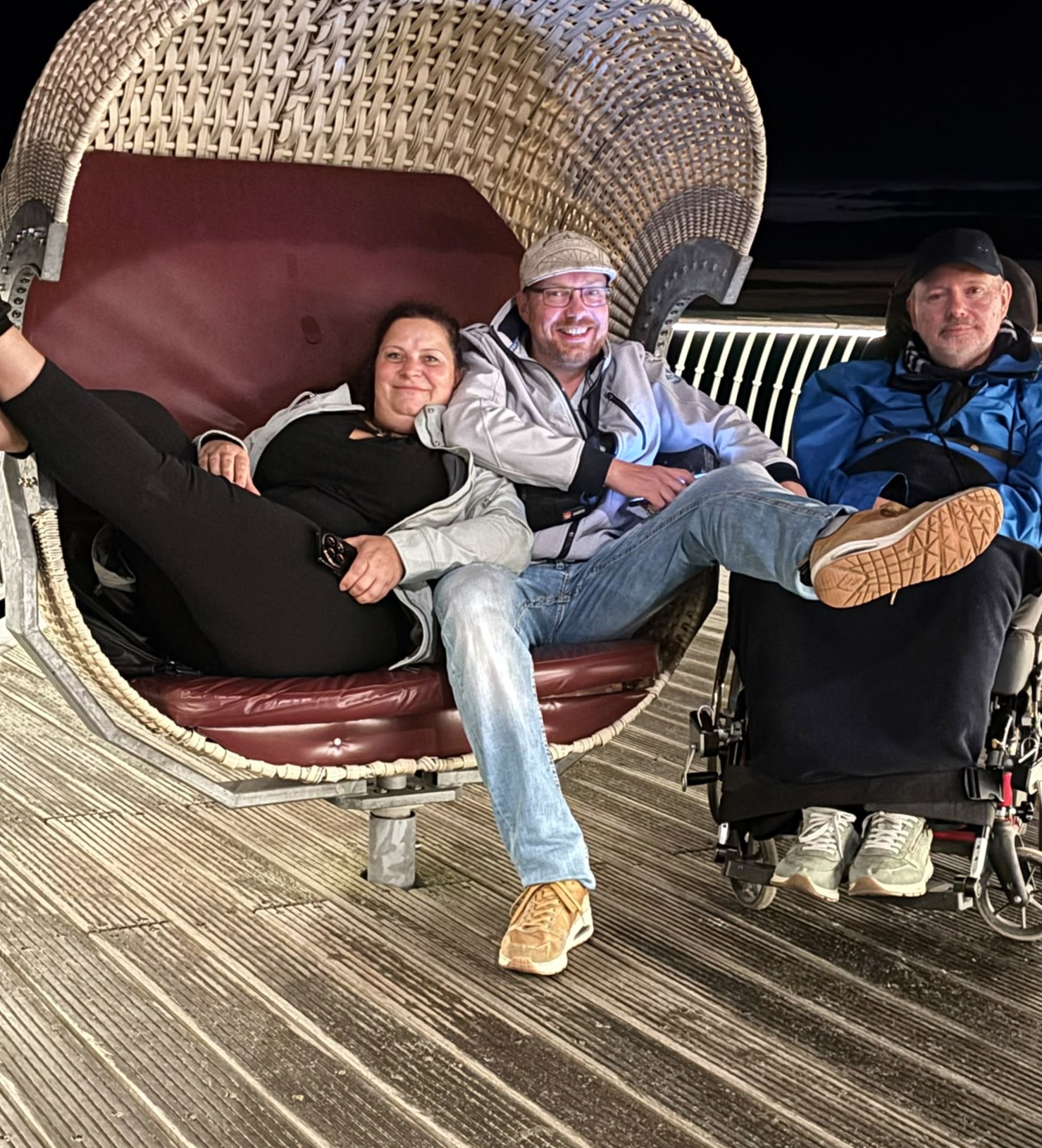 Drei Personen ruhen auf einem großen Korbstuhl auf einer Seebrücke