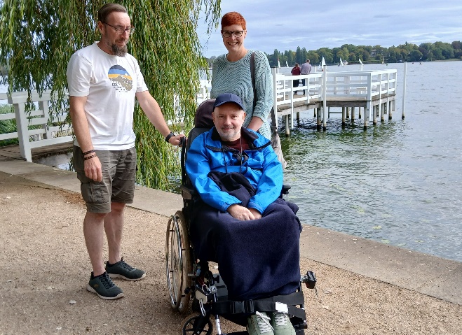Drei Personen am Ufer des Wannsee: ein Mann im Rollstuhl in blauer Jacke, dahinter ein Steg und Segelboote