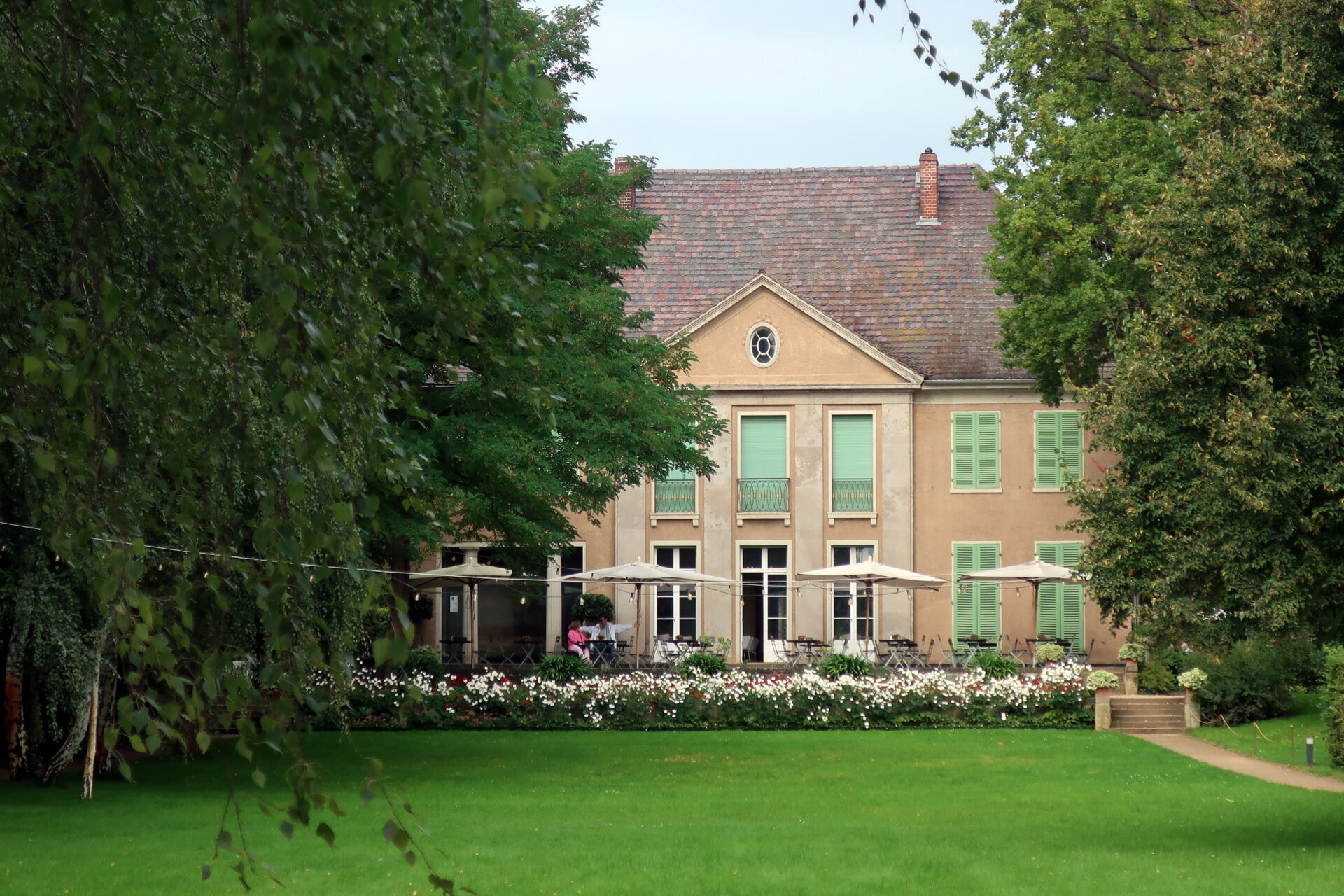 Die Liebermann-Villa von der Gartzenseite: zweistöckiges Haus mit Terrasse, im Garten sitzen Personen unter Sonnenschirmen